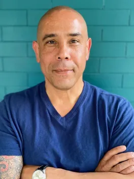A man in a dark blue short-sleeve shirt standing with arms crossed in front of a teal brick wall.